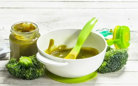 Babybrokkoli-Püree im Glas und Schüssel, daneben frischer Brokkoli und ein Löffel.