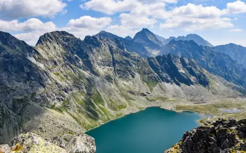 Widok na turkusowe jezioro u podnóża skalistych gór. W oddali widać kolejne pasma górskie, a na niebie chmury. To właśnie Koprowy Wierch.