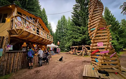Drewniany domek na drzewie i spiralna rzeźba z desek na festiwalu Grasz Bór. Ludzie spacerują, psy odpoczywają.