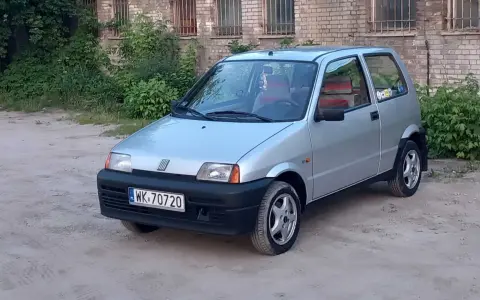 Srebrne cinquecento z czerwonymi akcentami we wnętrzu, zaparkowane na piaszczystym podwórku przed ceglanym budynkiem.