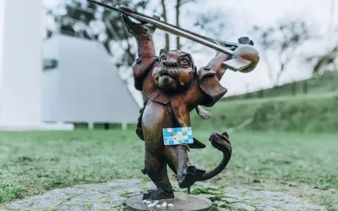 Figurka lew hewelion z kartą miejską w łapie, stojąca na bruku wśród zieleni.