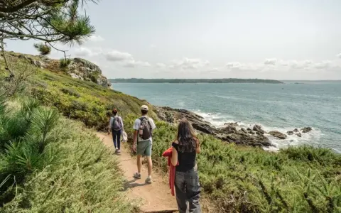 Trois randonneurs suivent un sentier côtier, profitant de la vue sur la mer. Ils avancent sur le chemin, comme dans les gr 34 étapes.
