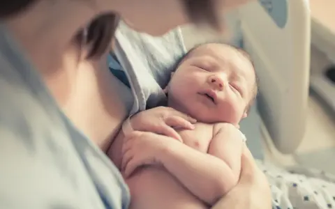 Noworodek śpi w ramionach matki, widoczna jej szyja i fragment ramienia.