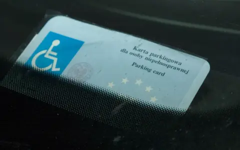 Niebieska karta parkingowa dla osoby niepełnosprawnej z symbolem wózka inwalidzkiego, umieszczona za przednią szybą samochodu.