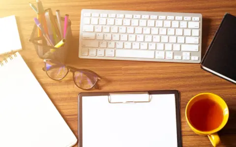 Schreibtisch mit Notizbuch, Stiften, Brille, Tastatur, auf der die tastatur y und z vertauscht sind, Klemmbrett und einer Tasse Tee.