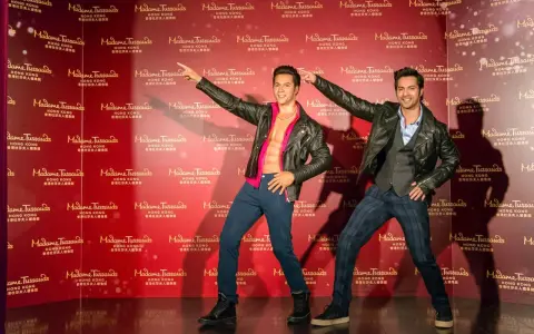 Dwie woskowe figury, jedna z odkrytym torsem, stoją na tle powtarzającego się logo Madame Tussauds w Hongkongu.