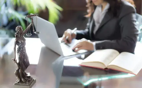 Kobieta w garniturze pracuje przy laptopie, obok Statua Temidy i otwarta księga. To droga, jak zostać prokuratorem.