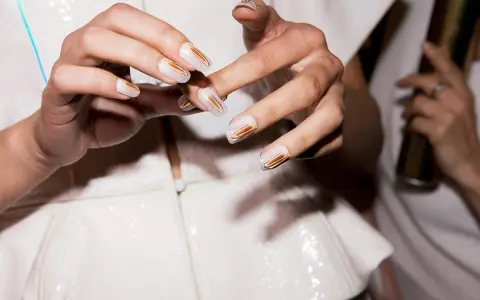 Eleganckie paznokcie przyklejane w białym kolorze ze złotymi zdobieniami. Dłonie prezentują nowoczesny manicure.