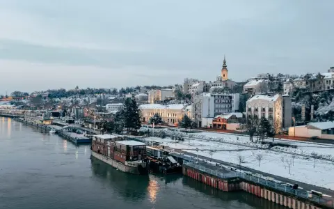 Widok na zimowe miasto nad rzeką z łodziami i ośnieżonymi budynkami.
