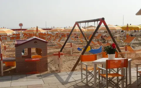 Area giochi e lettini sulla spiaggia 106 di Riccione, con ombrelloni arancioni e bianchi, altalena e casetta per bambini.