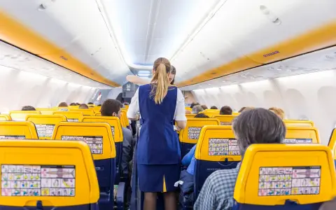 Wnętrze samolotu Ryanair, stewardessa obsługuje pasażerów. Żółte zagłówki siedzeń i pomarańczowe pasy na suficie tworzą słoneczną atmosferę.