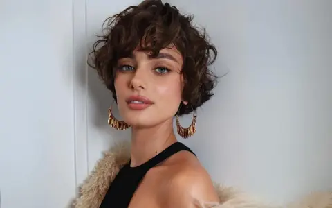 Mujer con un moderno corte shaggy rizado, ojos azules penetrantes y grandes pendientes dorados.