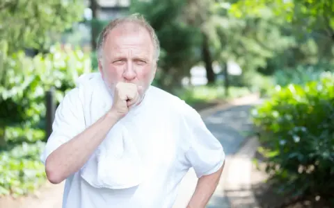 Mann mit COPD hustet im Freien. Sein dicker Bauch ist durch die weiße Kleidung sichtbar.