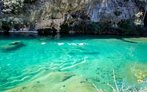 Persona nada en un pou clar de aguas turquesas, bajo un acantilado rocoso.