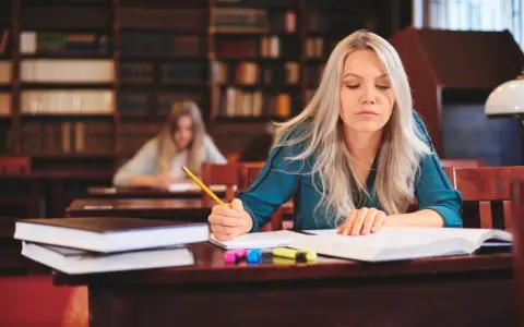 Kobieta z długimi blond włosami pisze w zeszycie, siedząc przy biurku w bibliotece.