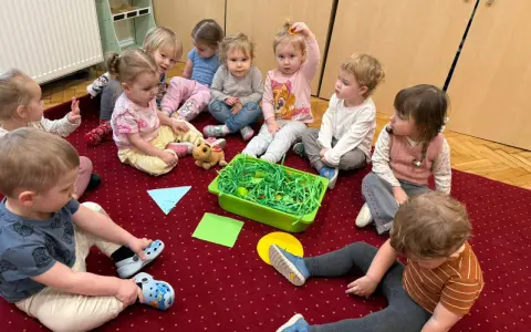 Dzieci siedzą na dywanie wokół zielonego pojemnika z zieloną trawą i jajkami.