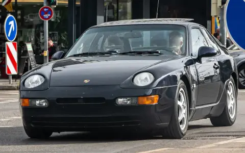 Dunkelblauer 968 Porsche fährt auf der Straße. Ein Mann sitzt am Steuer. Verkehrsschilder sind im Hintergrund zu sehen.