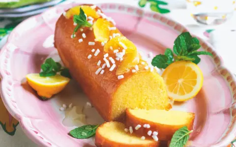 Torta al limone leggerissima, lucida e decorata con fettine d'arancia e zuccherini, servita su un piatto rosa con spicchi di limone e menta.