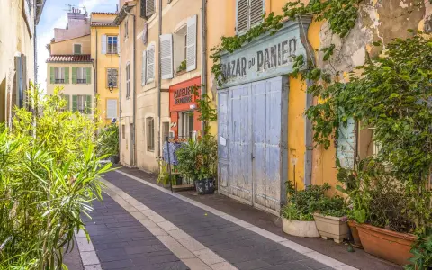 Bunte Häuser und Pflanzen säumen eine Gasse in der Marseille Altstadt. Ein "Bazar du Panier" Schild und blaue Holztüren fallen auf.