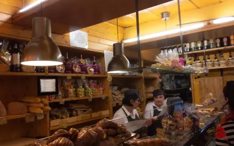 Interior of a bakery with shelves of bread, pastries, and bottles. Two women are behind the counter.