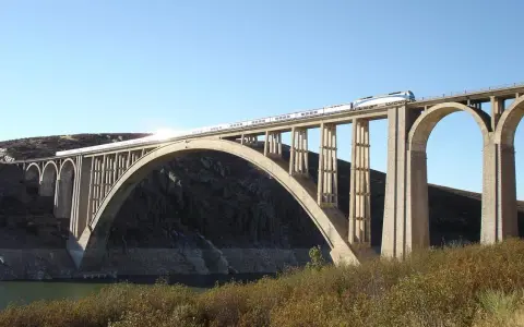 Un tren de alta velocidad cruza un imponente puente arco de hormigón sobre un embalse.