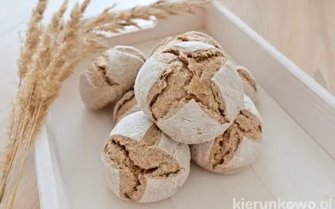 Kilka apetycznych bułek razowych na zakwasie, ułożonych na drewnianej tacy, obok ozdobnych traw.