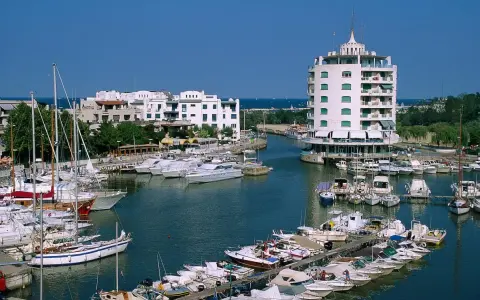 Un vivace porto verde con barche a vela e yacht ormeggiati, edifici bianchi e un edificio circolare a torre.
