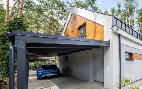 Nowoczesna wiata przy domu chroni niebieski samochód. Elegancka konstrukcja z ciemnym zadaszeniem i drewnianymi elementami architektonicznymi.