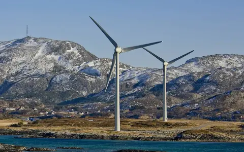 Zwei Windräder erzeugen sauberen Strom aus Norwegen. Im Hintergrund schneebedeckte Berge und ein kleines Dorf.