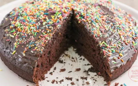 Ein saftiger, veganer Kuchen mit Schokoladenglasur und bunten Streuseln. Ein Stück fehlt, was Lust auf mehr macht.