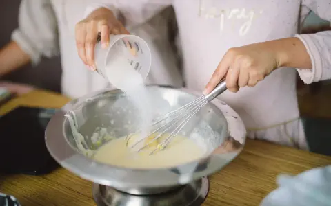 Préparation de la pâte à crêpes : on ajoute du sucre dans le bol avec un fouet. C'est la base de la recette des crêpes.