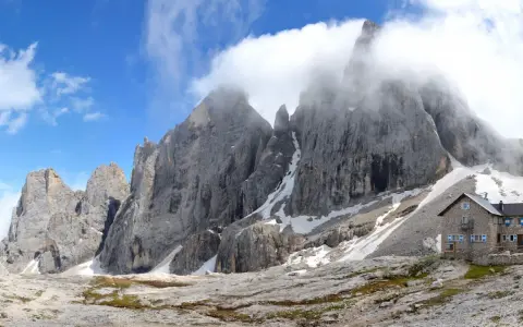 Rifugio in pietra ai piedi di imponenti vette rocciose, con neve residua e cielo azzurro. Un luogo incantevole al passo Valles.