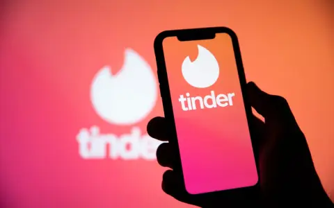 Dłoń trzyma smartfon z logo Tinder. W tle widać większe, rozmyte logo aplikacji, sugerujące nowe tinder powiadomienia.