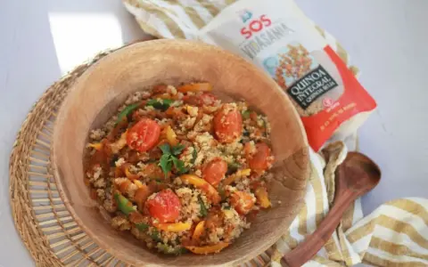 Un tazón de madera lleno de quinoa con verduras coloridas: tomates cherry, pimientos verdes y amarillos. Cerca, un paquete de quinoa integral y roja.