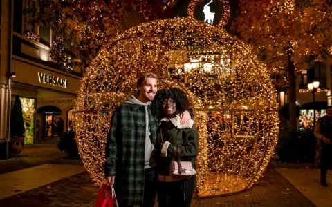 Ein glückliches Paar vor einer riesigen Lichterkugel auf dem weihnachtsmarkt roermond. Geschäfte wie Versace und Ralph Lauren sind im Hintergrund zu sehen.