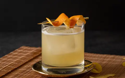 Ein erfrischender Rum Sour mit Eis und Orangenzesten garniert, serviert auf einem Bambusuntersetzer.