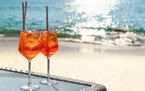 Zwei Aperol Spritz Gläser mit Eis und Orangenscheiben stehen am Strand. So trinkt man Aperol Spritz in Italien!