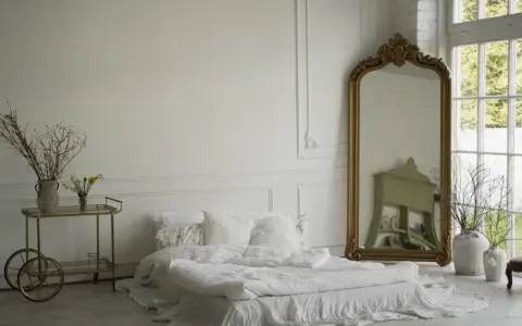 Elegancka sypialnia z łóżkiem w bieli, ozdobiona sztukaterią przypominającą lamperię angielską. Złote lustro i wózek dodają blasku.