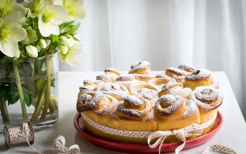 Torta di rose soffice e dorata, cosparsa di zucchero a velo, decorata con un nastro a cuoricini. Accanto, un vaso con fiori bianchi.
