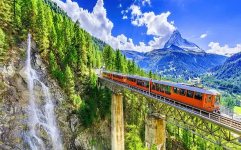 An orange train crosses a bridge over a waterfall, on its way from Zurich to Andermatt. Lush green trees and mountains surround the scenic route.