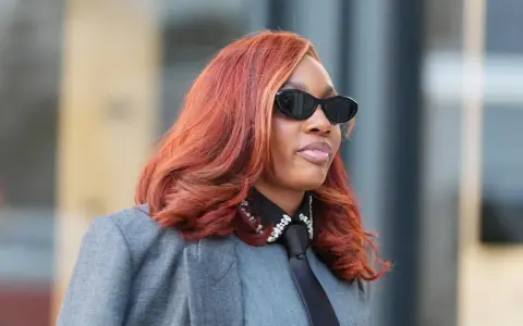Frau mit leuchtend roten Haaren, Sonnenbrille und grauem Blazer.