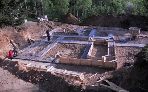 Budowa fundamentów: zbrojenie i szalunki czekają na chudy beton. W tle las i plac budowy.