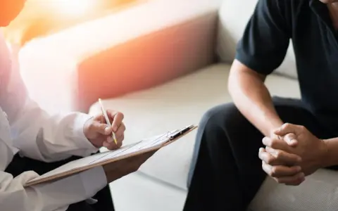 En terapeut skriver anteckningar medan en patient sitter med händerna knäppta. Bilden illustrerar vad betyder terapi – en plats för samtal och stöd.