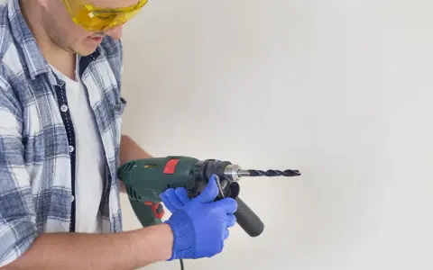 Man in safety glasses and blue gloves holding a drill to a white surface.