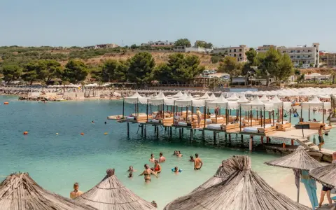 Słoneczny dzień na Ksamil Beach. Ludzie kąpią się w turkusowej wodzie, relaksują się na leżakach pod białymi parasolami i na drewnianych pomostach.