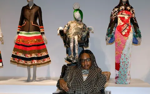 Bei einer Vernissage werden extravagante Kleider auf Schaufensterpuppen präsentiert. Eine Frau im Rollstuhl sitzt davor.