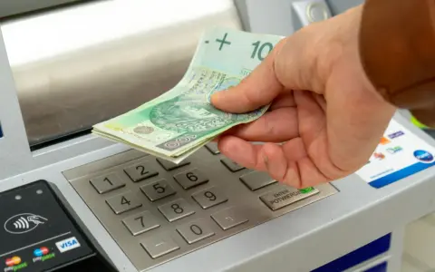 Dłoń wkłada banknoty 100 zł do bankomatu. Zastanawiasz się, ile można wpłacić na konto?