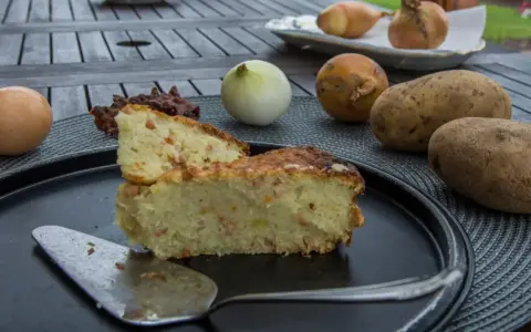 Babka ziemniaczana przepis babci: złocisty placek z ziemniaków, jajek i boczku, obok surowe ziemniaki, cebula i jajko.