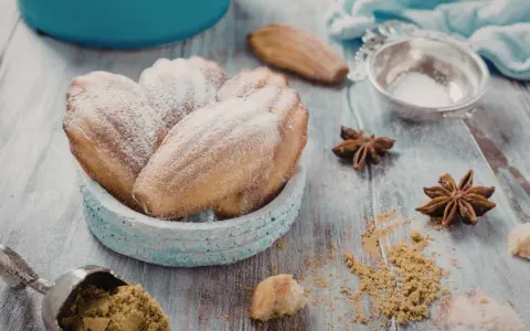 Un bol de biscuits madeleine noisette saupoudrés de sucre glace, avec des étoiles de badiane et une cuillère de poudre sur une table en bois.