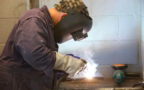 Soldador trabajando con un electrodo, creando chispas y humo. Se aprecian partes del electrodo en acción.